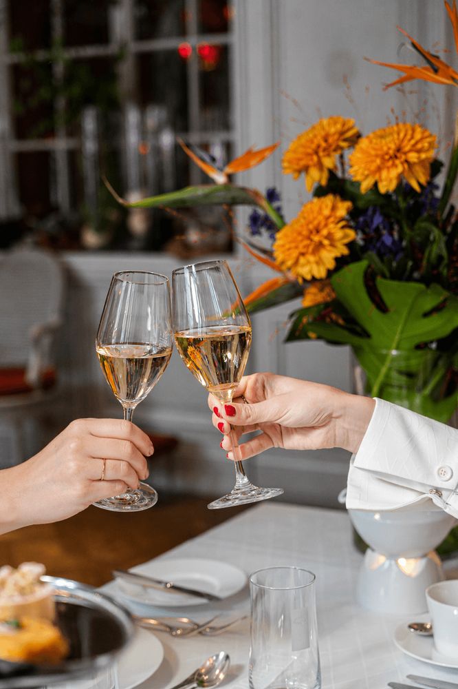 Zwei Personen stoßen mit Gläsern Weißwein an einem gedeckten Tisch mit Blumen zur Dekoration an.