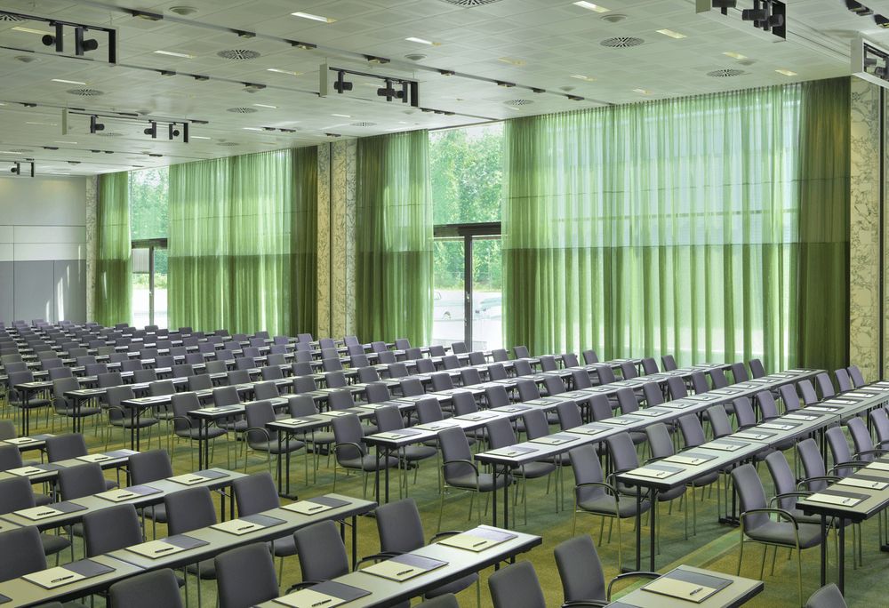 A large, empty conference or lecture room with rows of tables and chairs, and green curtains covering the windows.