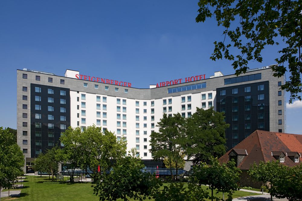 Vícepodlažní budova hotelu s nápisem 'Steigenberger Airport Hotel' na střeše, obklopená stromy a…