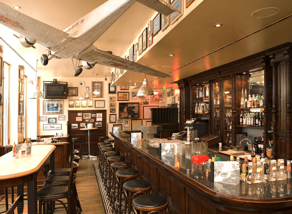 Das Foto zeigt das Innere einer Bar oder eines Pubs mit einer langen Theke, Barhockern, Tischen, Stühlen und einem Modellflugzeug, das von der Decke hängt.