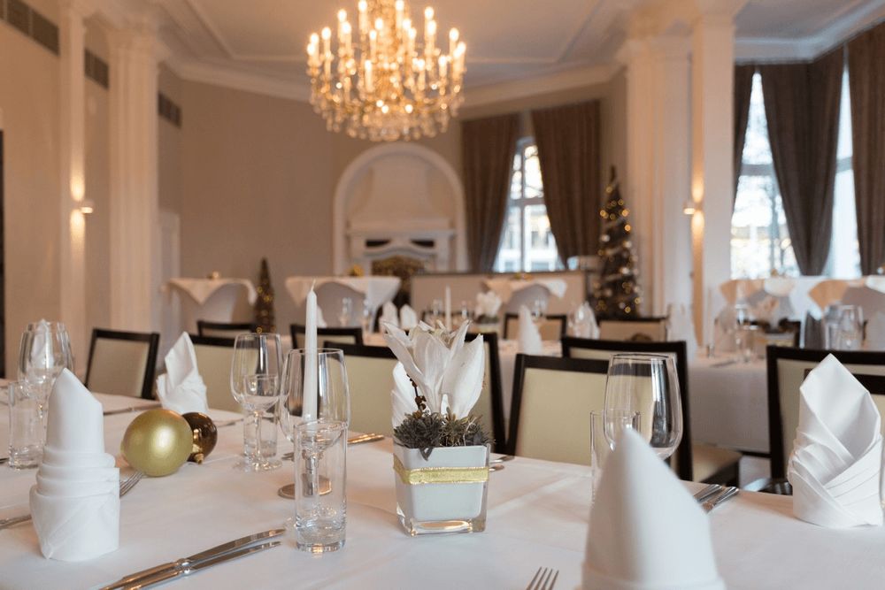 A foto mostra uma sala de jantar formal montada com mesas, toalhas brancas, guardanapos dobrados,…