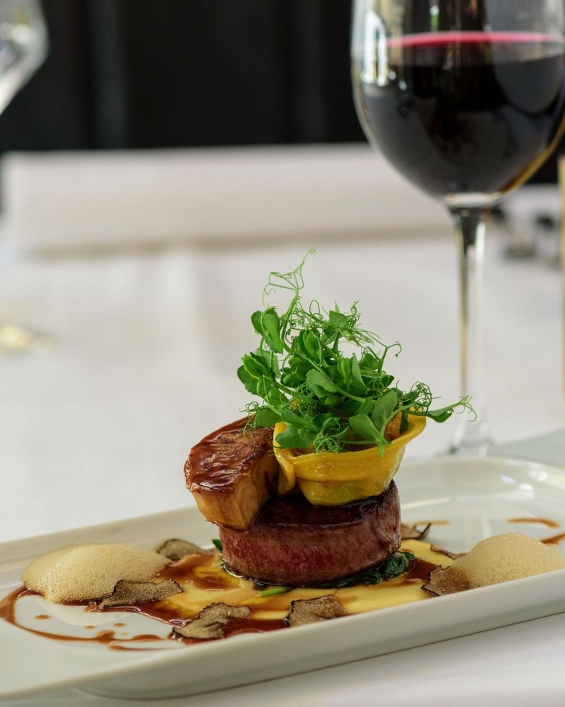 A plated gourmet dish with a piece of meat topped with greens and sauce, accompanied by a glass of…