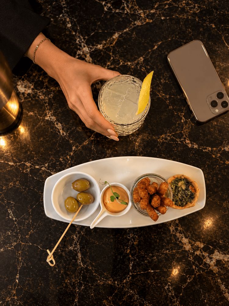 A hand holding a drink is shown above a black marble table with a smartphone and a dish containing…