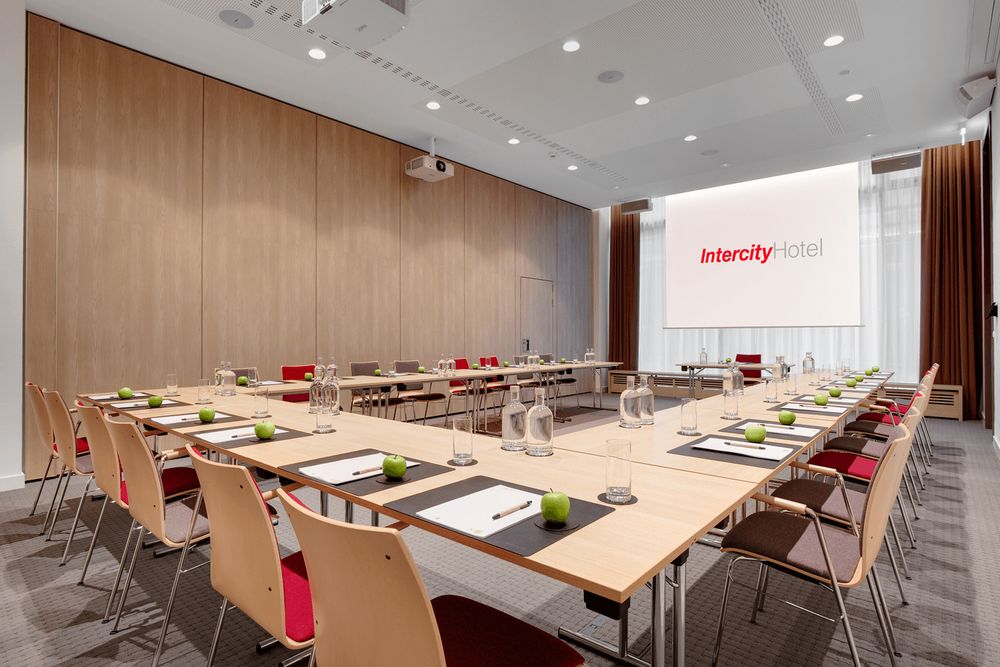 A conference room set up with tables arranged in a U-shape, equipped with chairs, notepads, pens, water glasses, water pitchers, and green apples. A projector screen at the front displays the logo "Intercity Hotel."