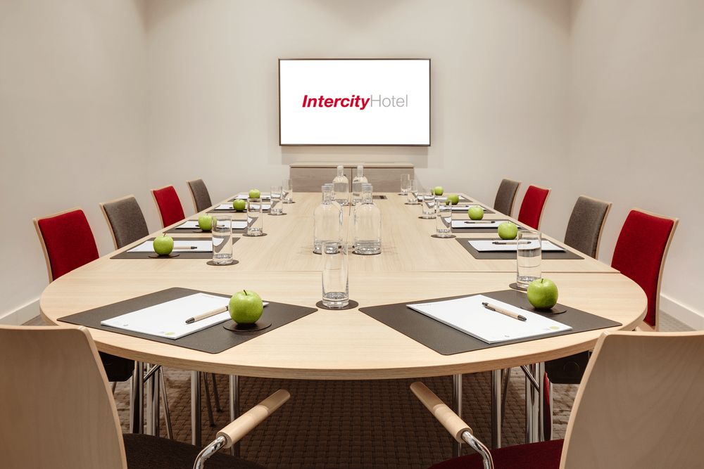 A conference room set up with a large wooden table, chairs, notepads, pens, green apples, water glasses, and bottles, with a screen displaying the Intercity Hotel logo.