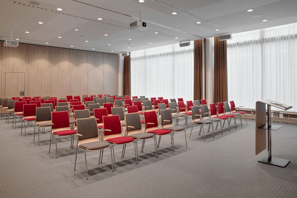 A modern conference room with rows of red and gray chairs arranged facing a podium and a projector.