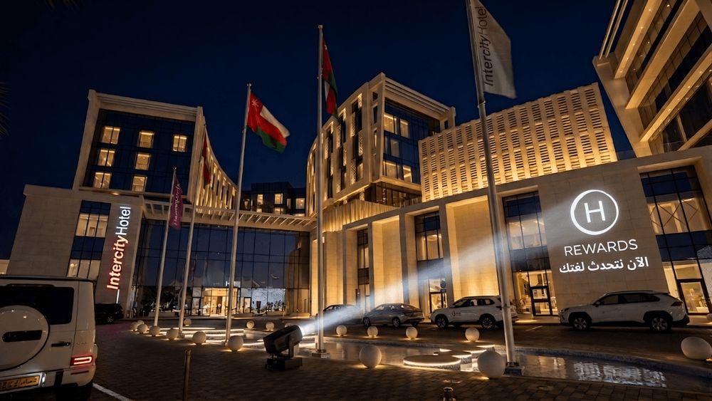 Ein modernes Hotelfgebäude bei Nacht mit beleuchteten Fahnen und geparkten Autos davor.