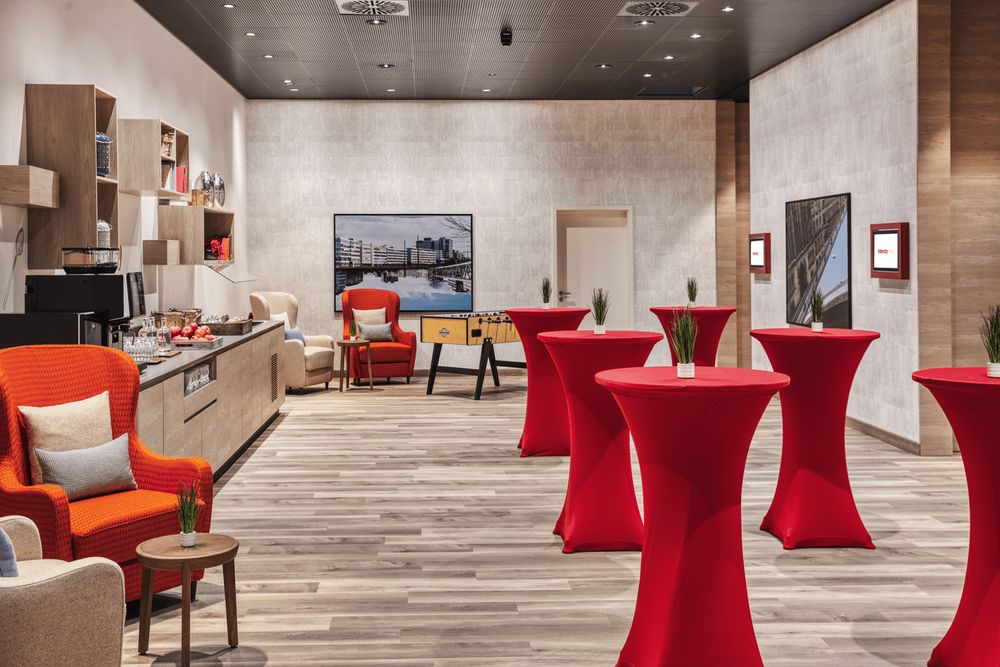 The photo shows a modern lounge area with high tables covered in red cloths, chairs, and a kitchenette in the background.