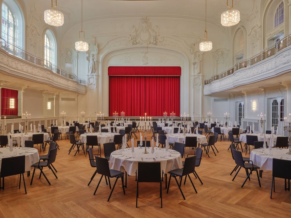 Ein großer eleganter Bankettsaal mit runden Tischen und Stühlen, einer Bühne mit rotem Vorhang…