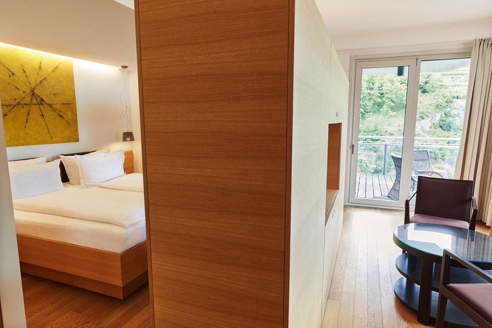 Una habitación de hotel moderna con un área de cama en un lado y un área de estar con balcón en el otro, separadas por un tabique de madera.