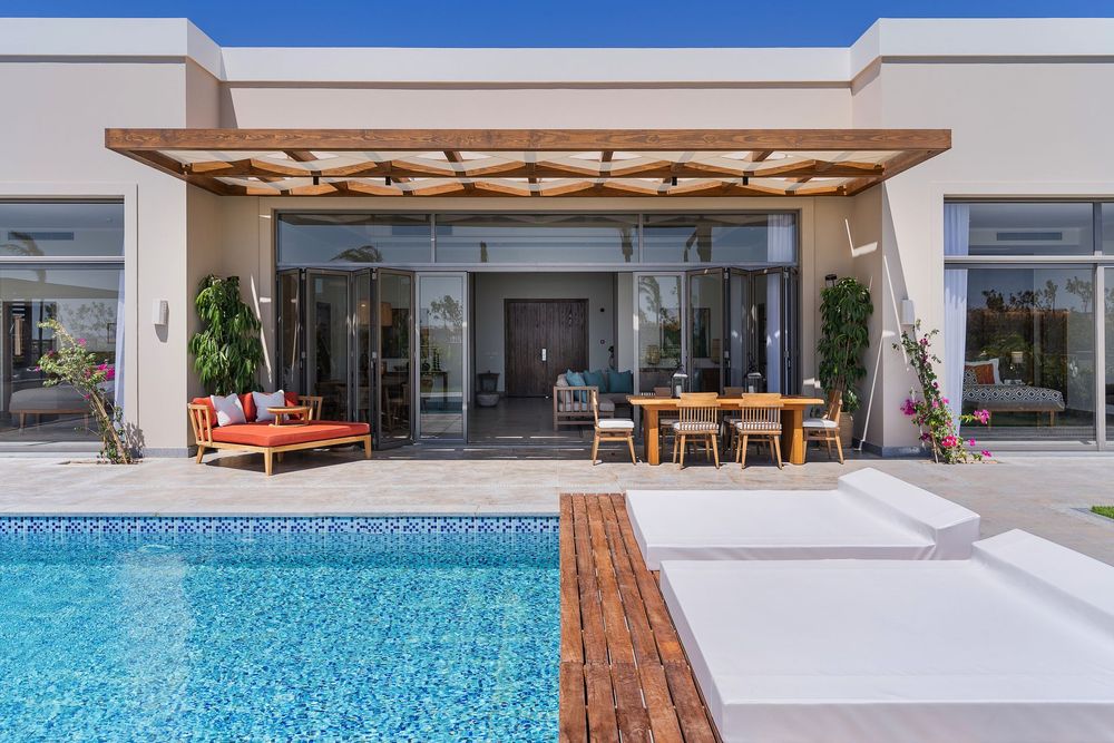 تُظهر الصورة منطقة فناء خارجية حديثة مع مسبح، وطاولة خشبية، وكراسي استلقاء، وبيرغولا، وأثاث جلوس…