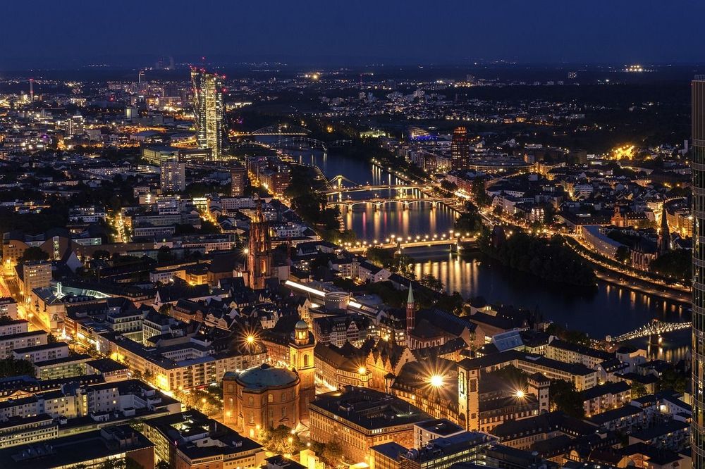 A nighttime aerial view of a city with illuminated buildings and bridges reflecting on a river.