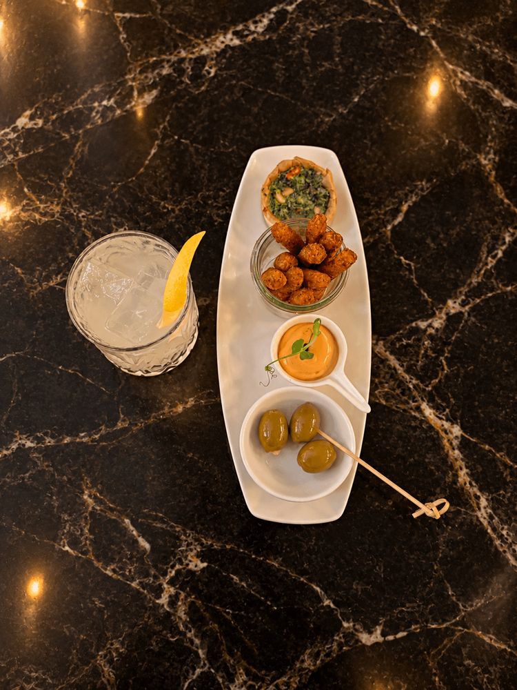A beverage with a lemon peel garnish next to a serving plate containing olives, a creamy dip,…