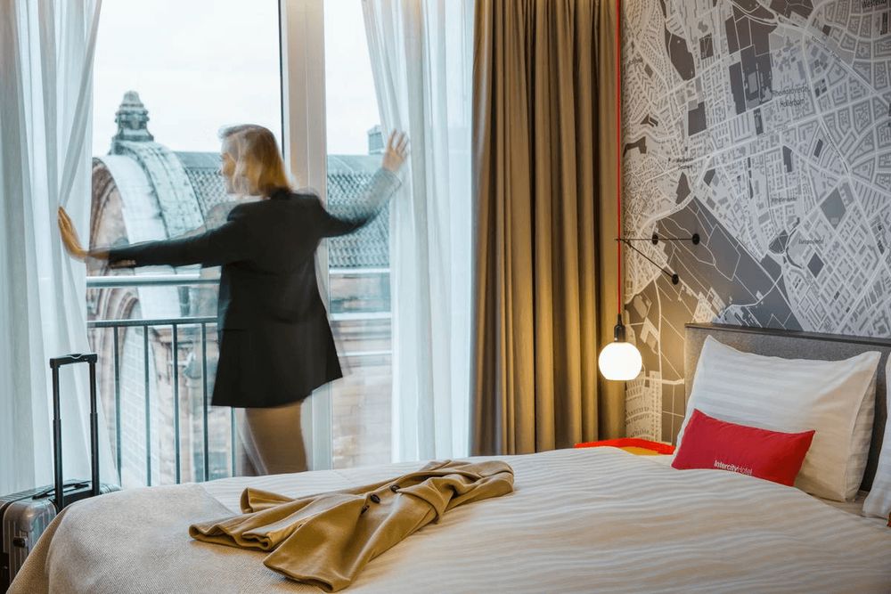 Eine Person steht an einem Fenster in einem Hotelzimmer, mit einem Koffer in der Nähe und einem…