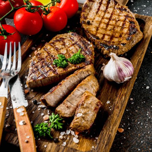 Ein Holzbrett mit gegrillten Steaks, Tomaten, Knoblauch und einigen Kräutern, begleitet von Messer und Gabel.