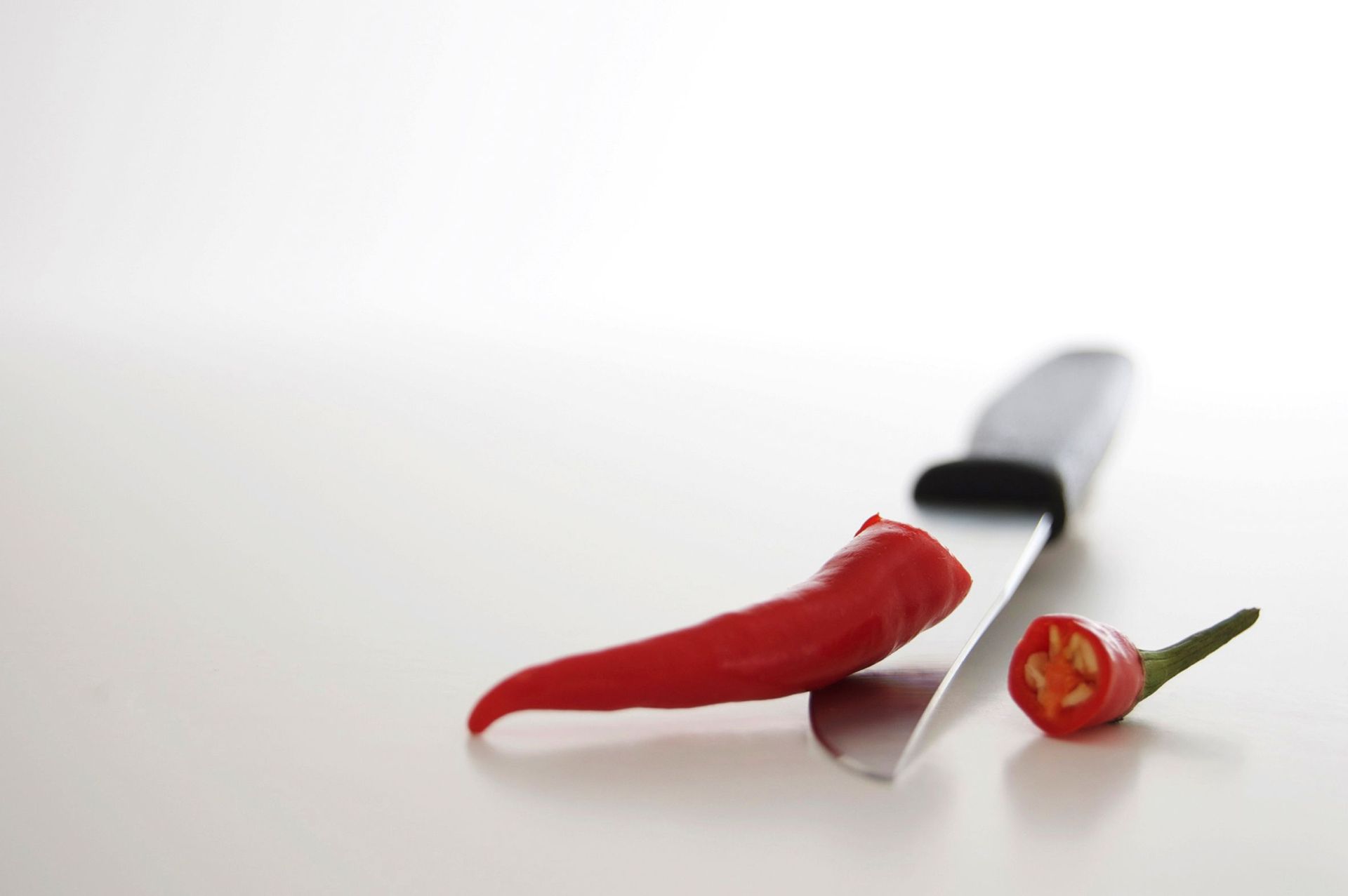 Das Foto zeigt eine rote Chilischote, von der eine Hälfte geschnitten ist, neben einem Messer auf…