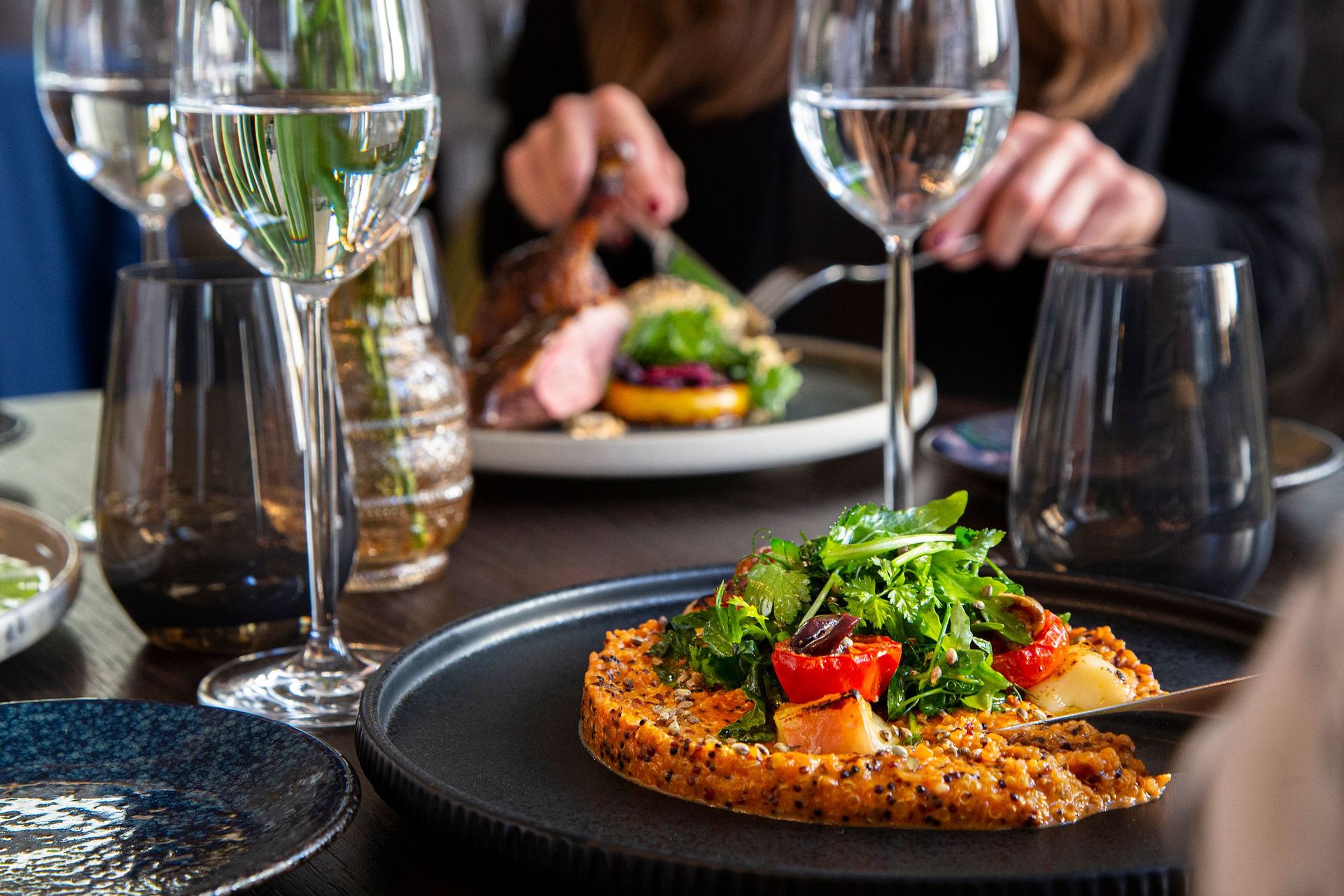 Eine Nahaufnahme eines Esstisches mit Essen, Gläsern mit Wasser oder Wein und einer Person, die im…