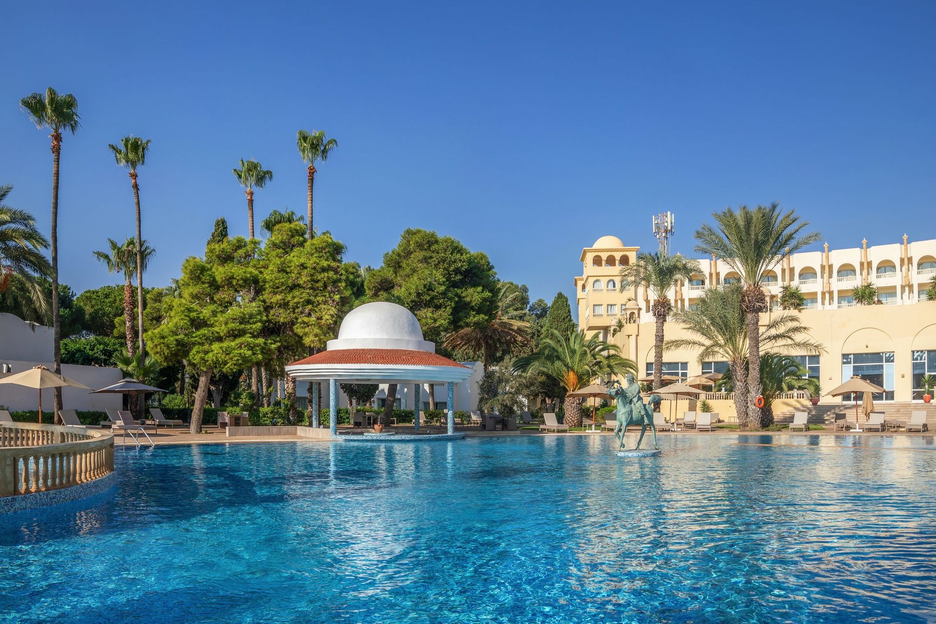 A large outdoor swimming pool with surrounding palm trees and a building in the background.