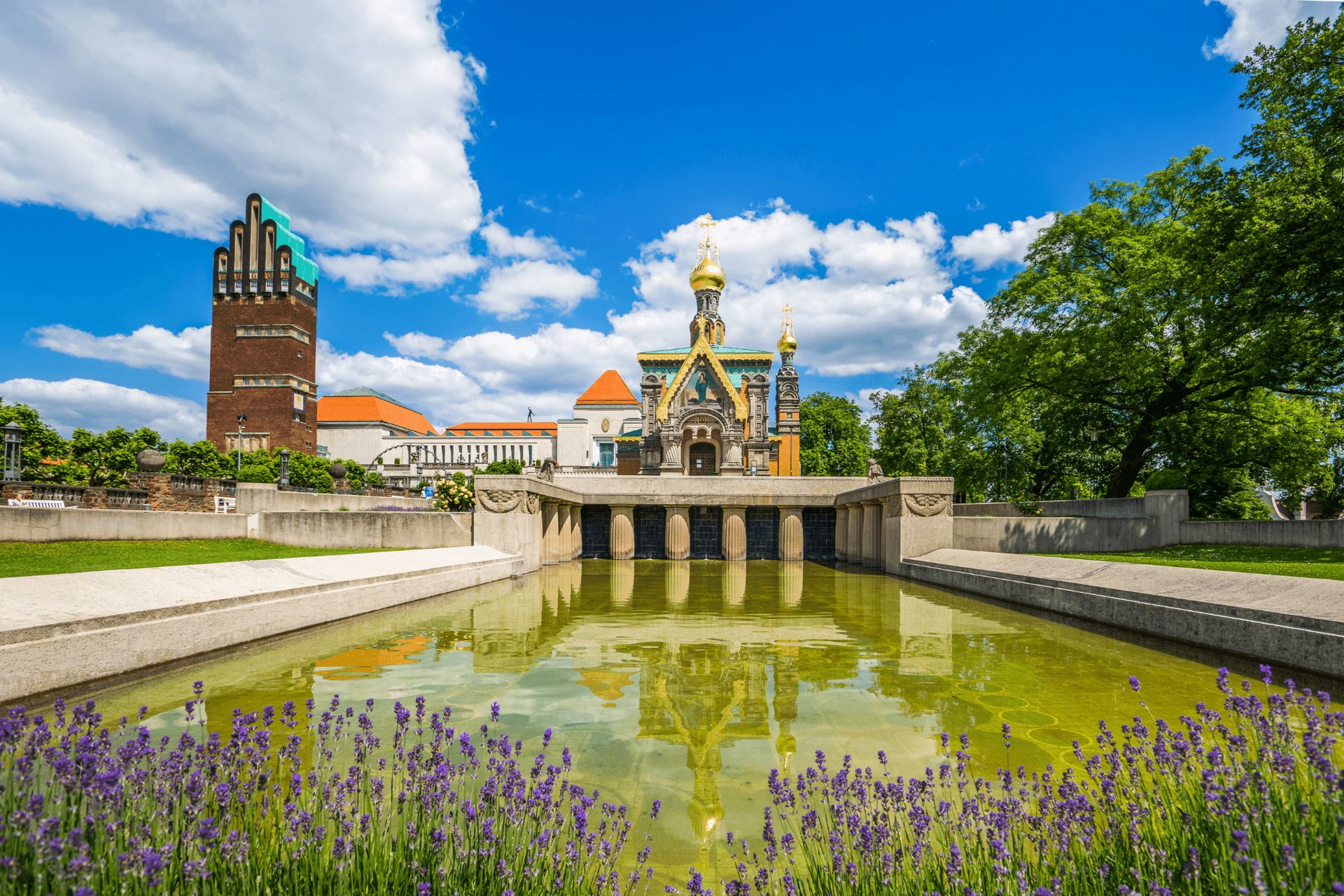 A historic building and a modern tower are shown with a reflective pond and purple flowers in the…