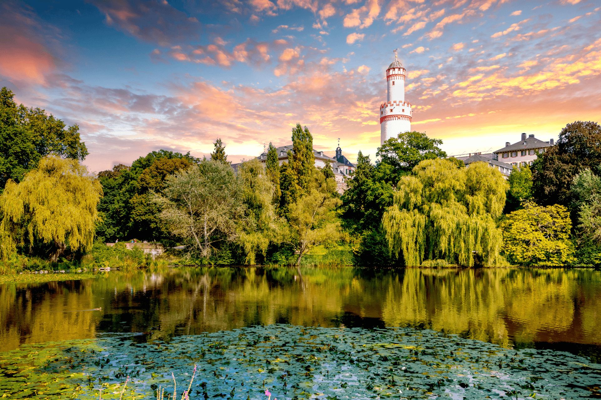 A scenic view of a park with a pond in the foreground, lush green trees, and a tower in the…