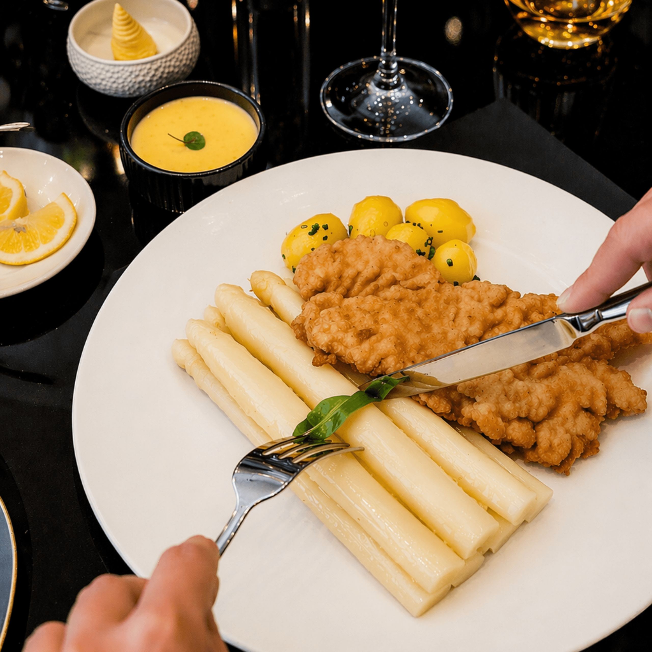 Ein Teller mit paniertem Fleisch, weißem Spargel und kleinen Kartoffeln wird mit Gabel und Messer…