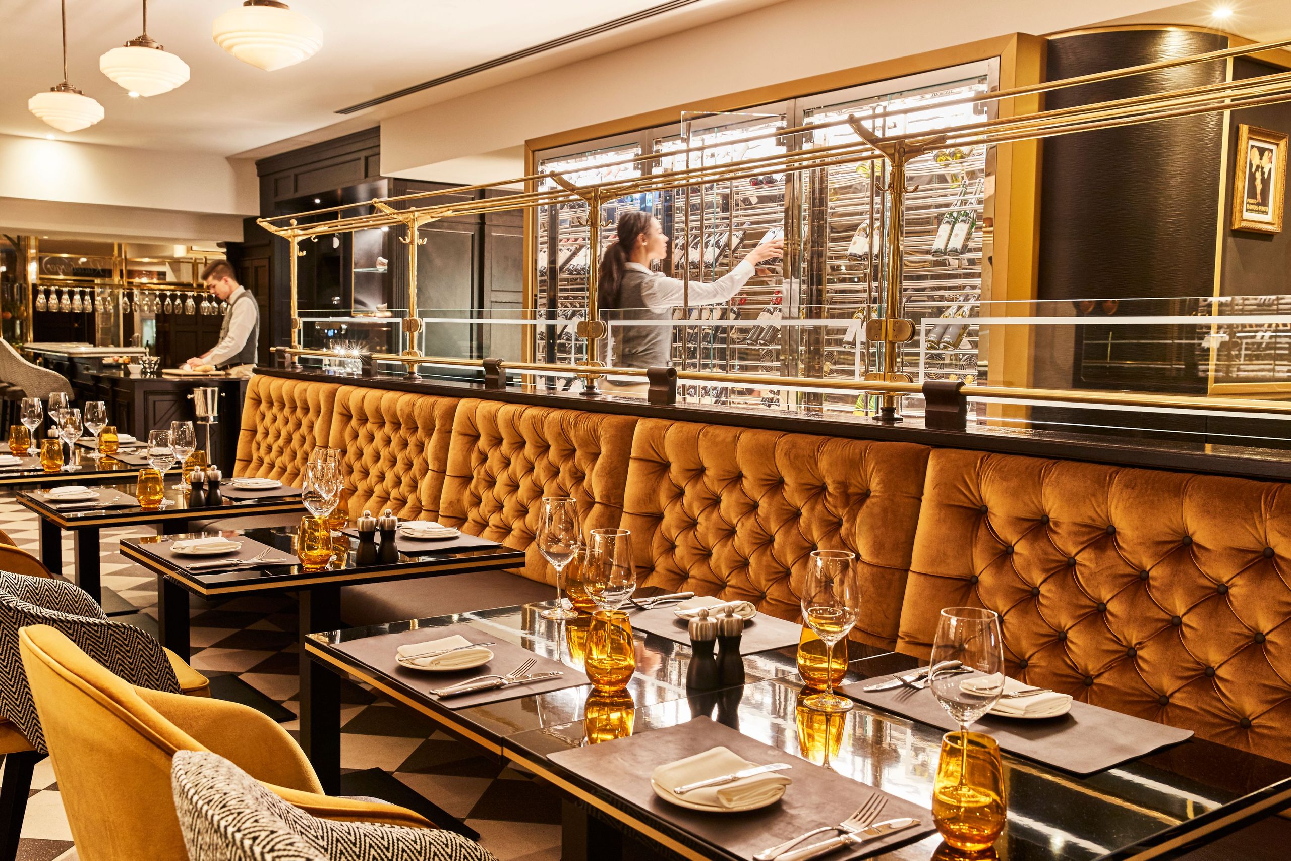 The photo shows the interior of an elegant restaurant with neatly set tables and two staff members…