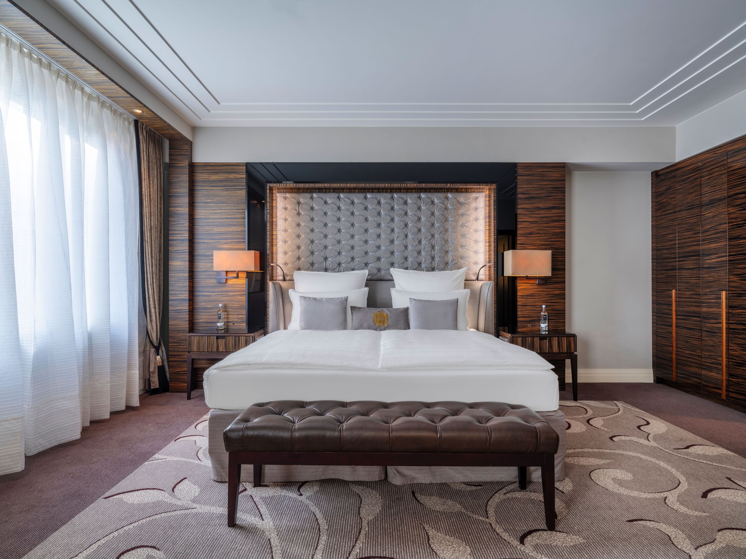 A modern and elegant bedroom with a large bed, bedside tables with lamps, a bench at the foot of…