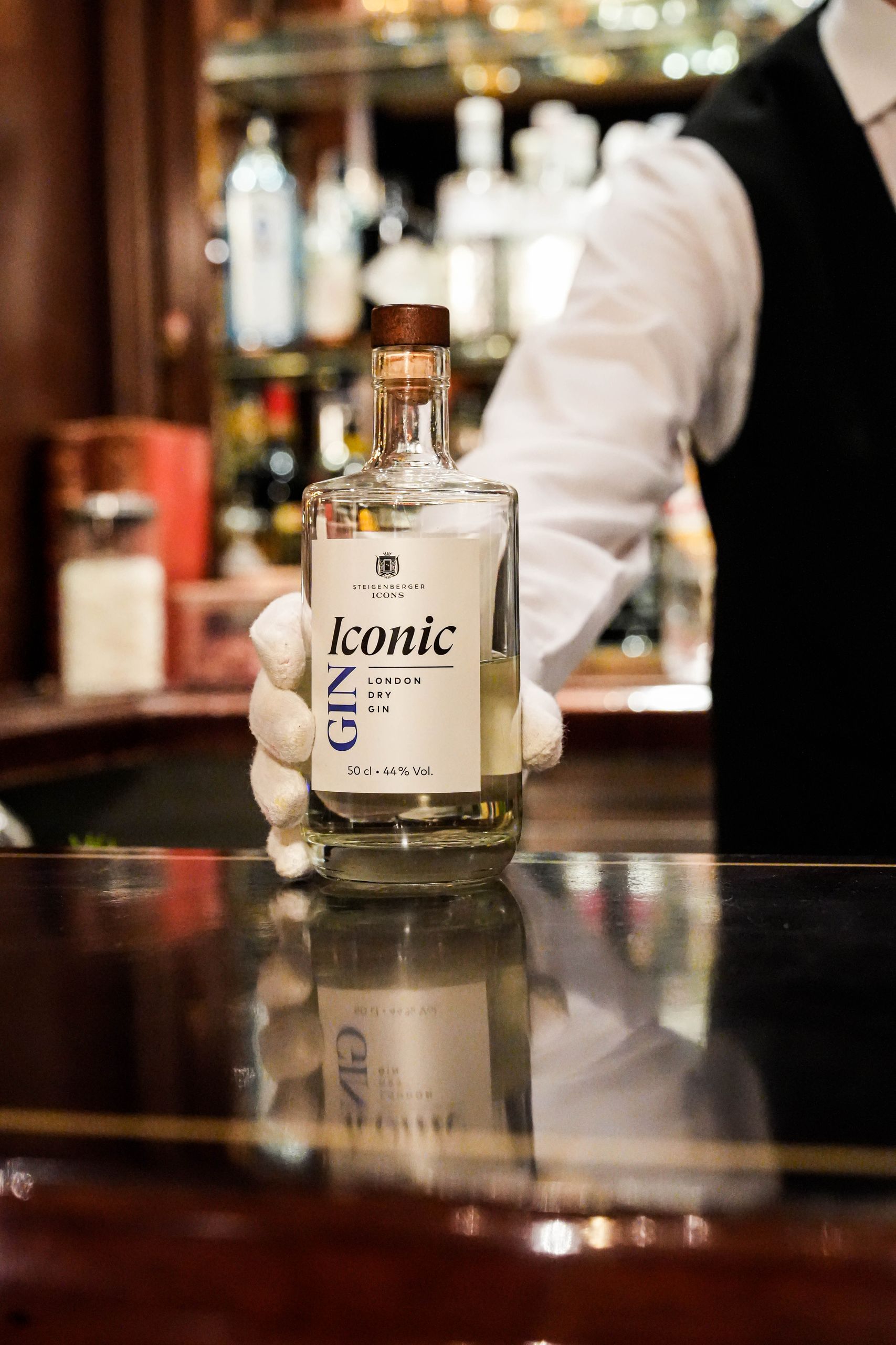 A person in a white shirt and black vest is holding a bottle of Iconic London Dry Gin.