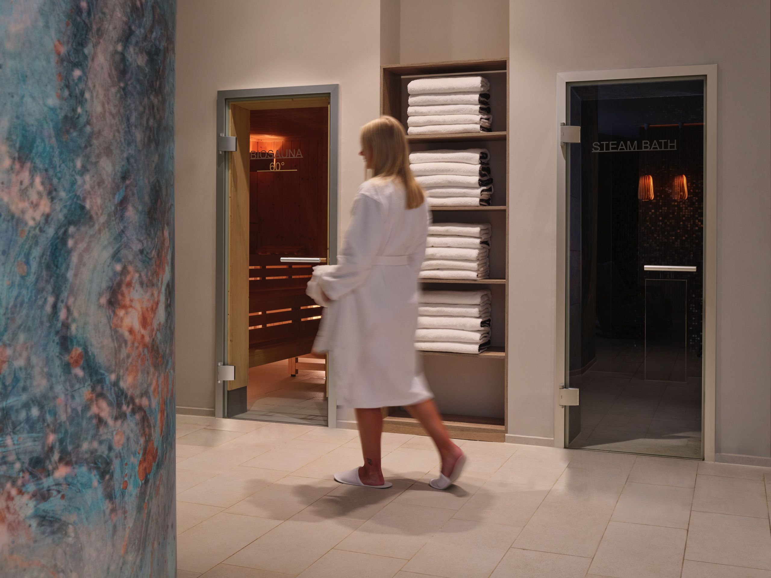 Eine Person in einem weißen Bademantel geht in einem Spa- oder Wellnessbereich an Räumen vorbei,…
