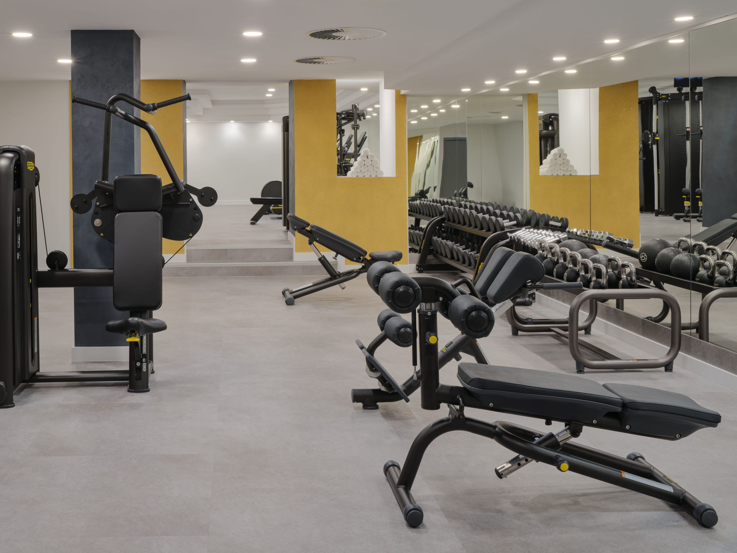 Das Foto zeigt ein Fitnessstudio mit verschiedenen Trainingsgeräten, darunter Bänke,…