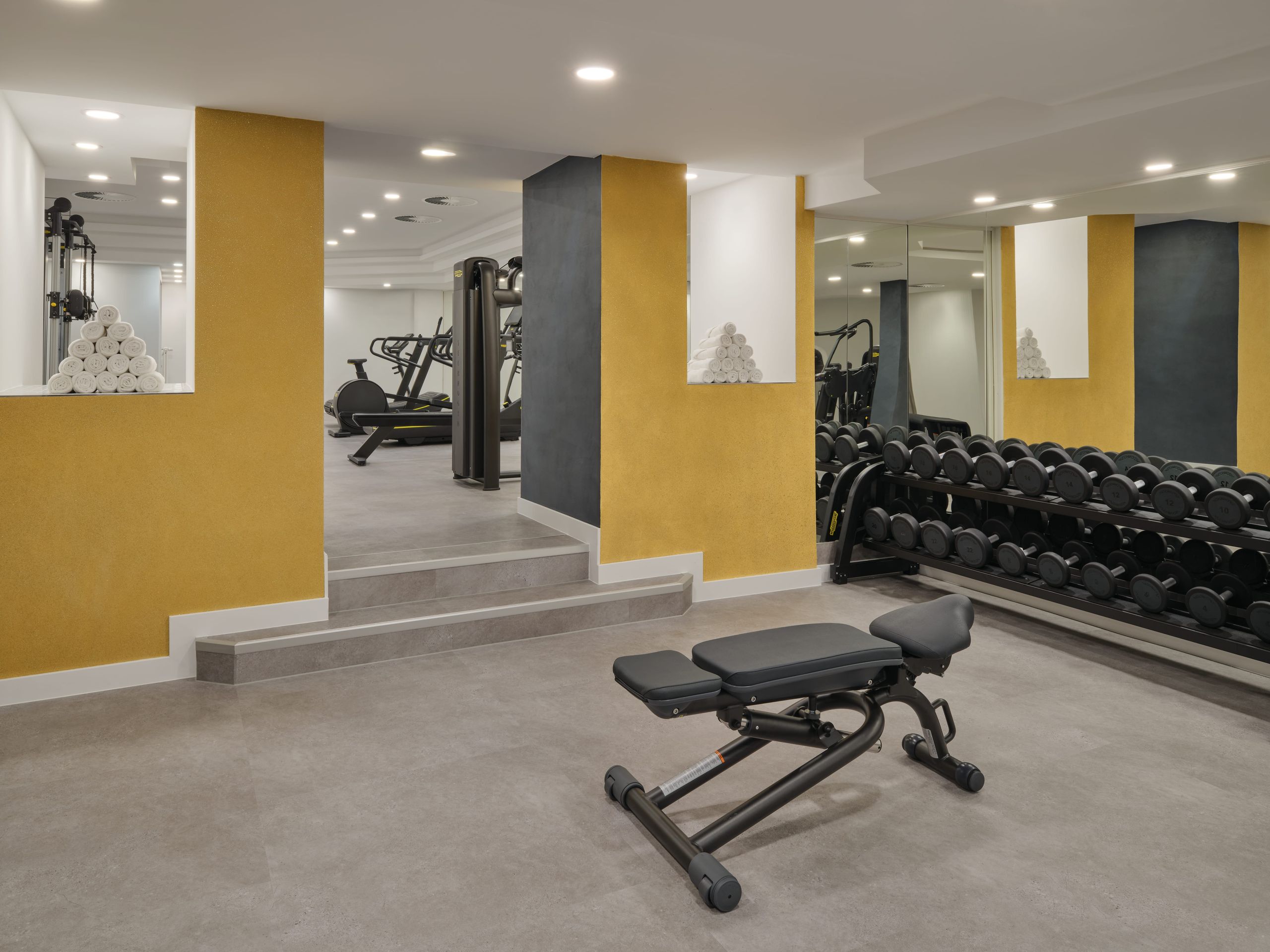 Das Foto zeigt ein Fitnessstudio mit Trainingsgeräten wie einer Trainingsbank, Kurzhanteln und…