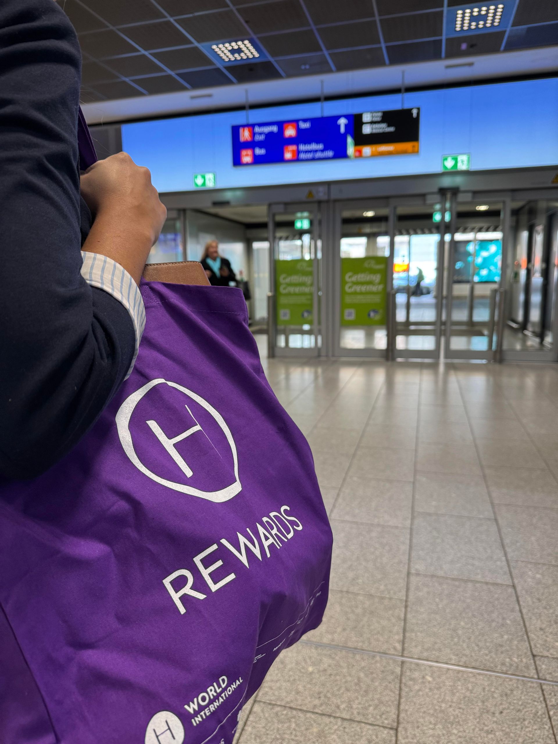 A person holding a purple tote bag standing inside an airport terminal near the entrance or exit…