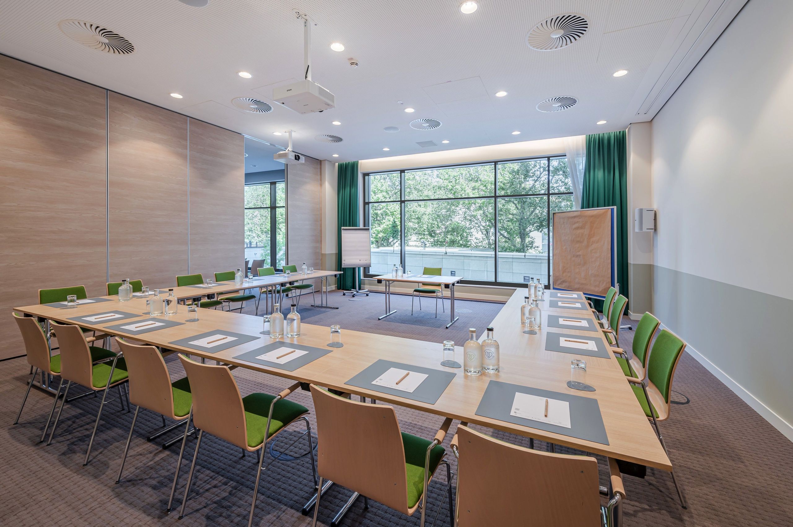 A modern conference room with a U-shaped table setup, chairs, notepads, water bottles, and large…