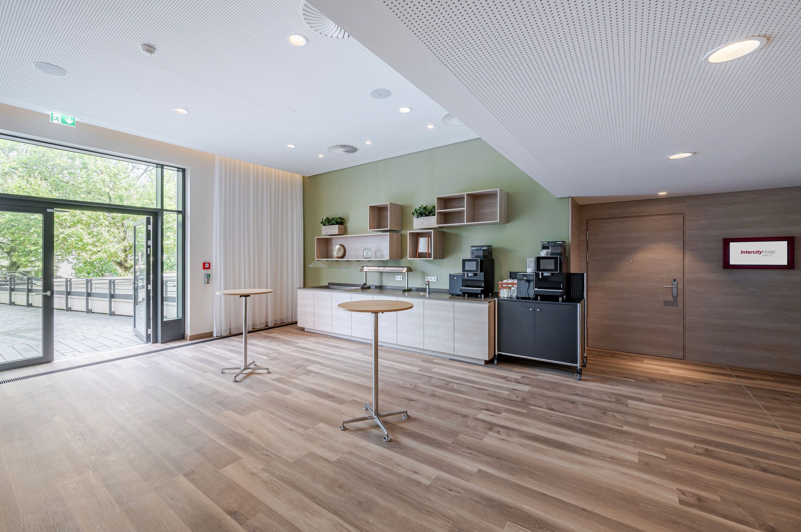 The photo shows a modern, spacious room with a coffee station, tall tables, wooden flooring, and…