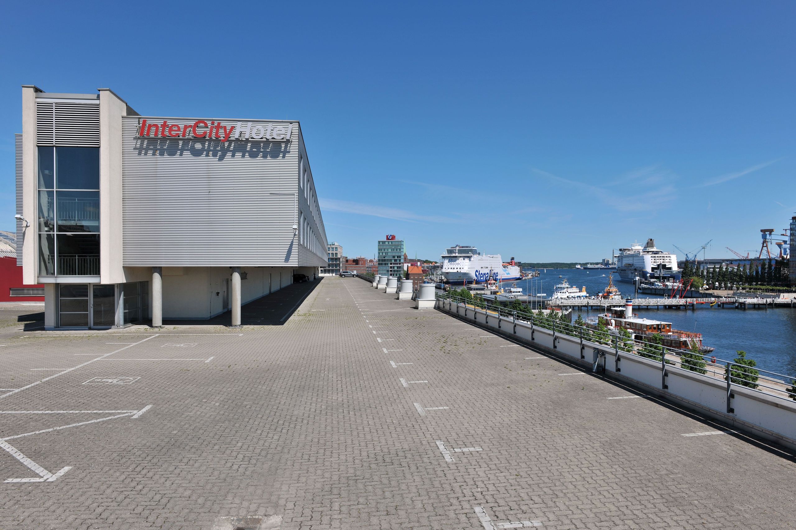 Uma área à beira-mar com um edifício de hotel à esquerda e várias embarcações atracadas à direita…