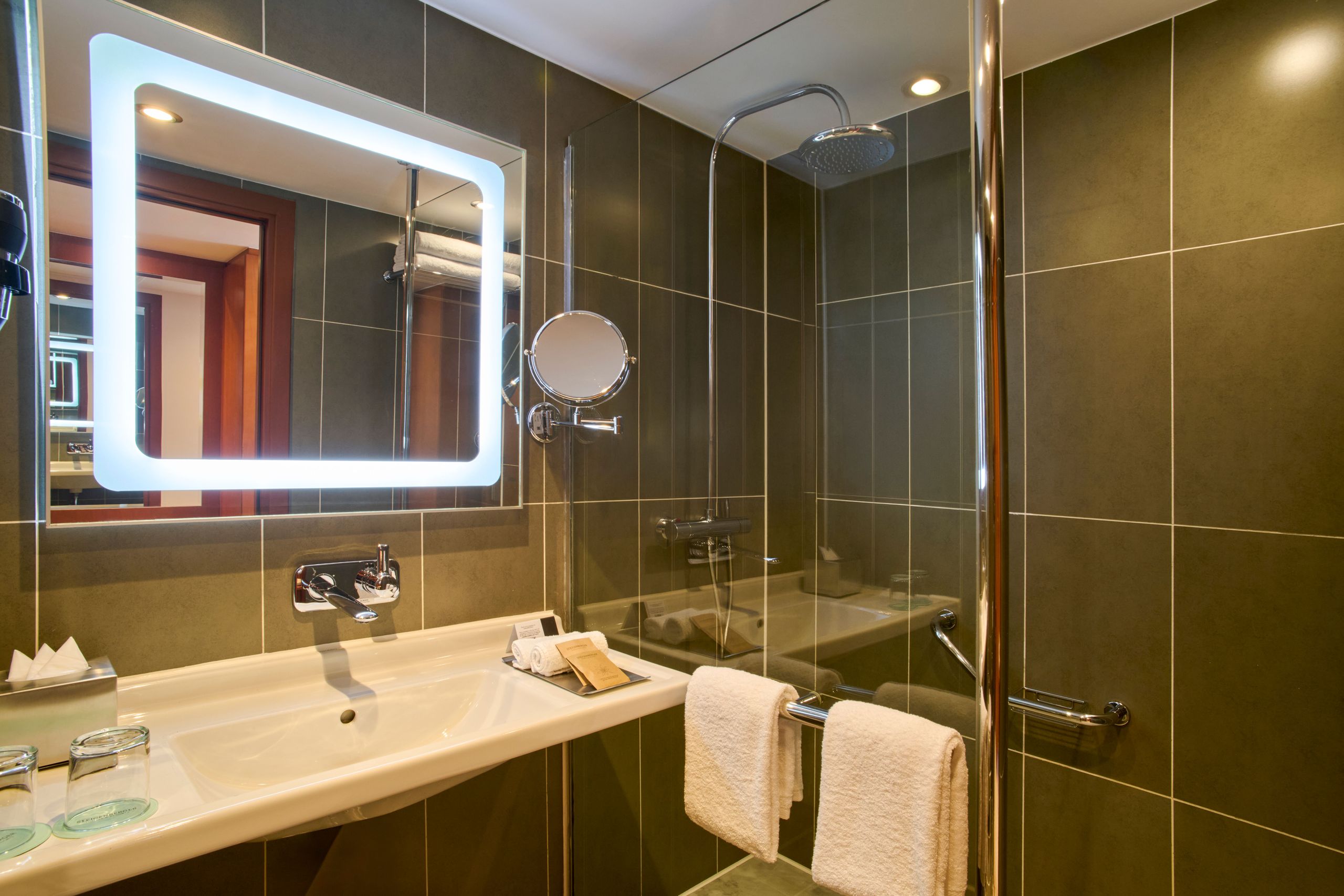 La photo montre une salle de bain moderne avec un lavabo, un miroir éclairé et une douche vitrée…