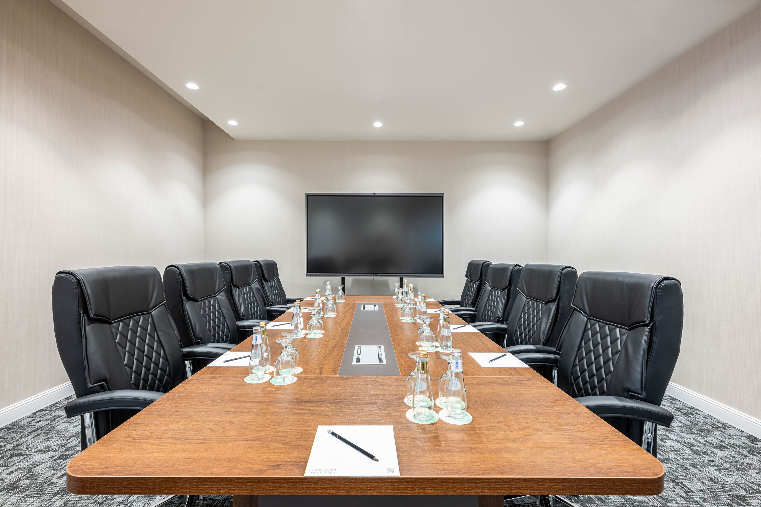 A conference room with a long wooden table, black chairs, and some stationery and glasses arranged…