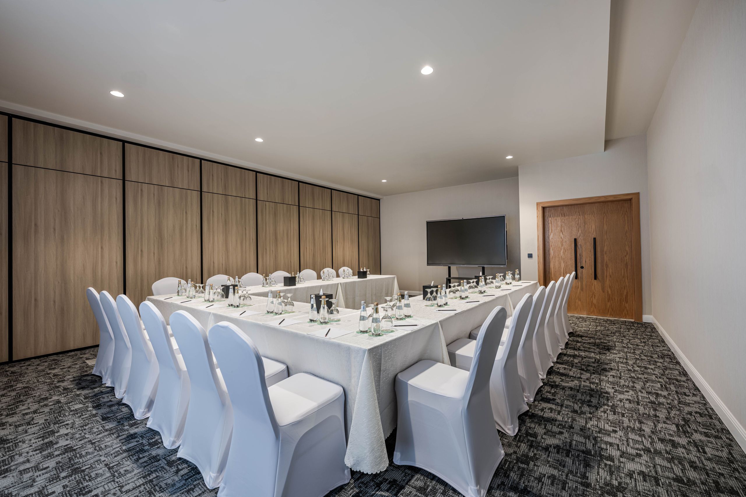 A conference room set up with tables arranged in a U-shape, surrounded by white-covered chairs,…