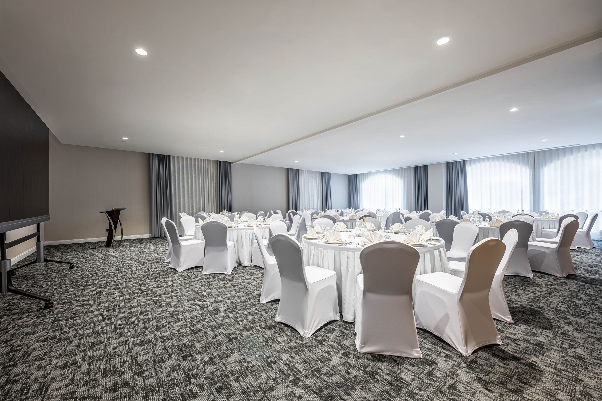 A spacious banquet hall with round tables and chairs covered in white cloth, arranged for an event…