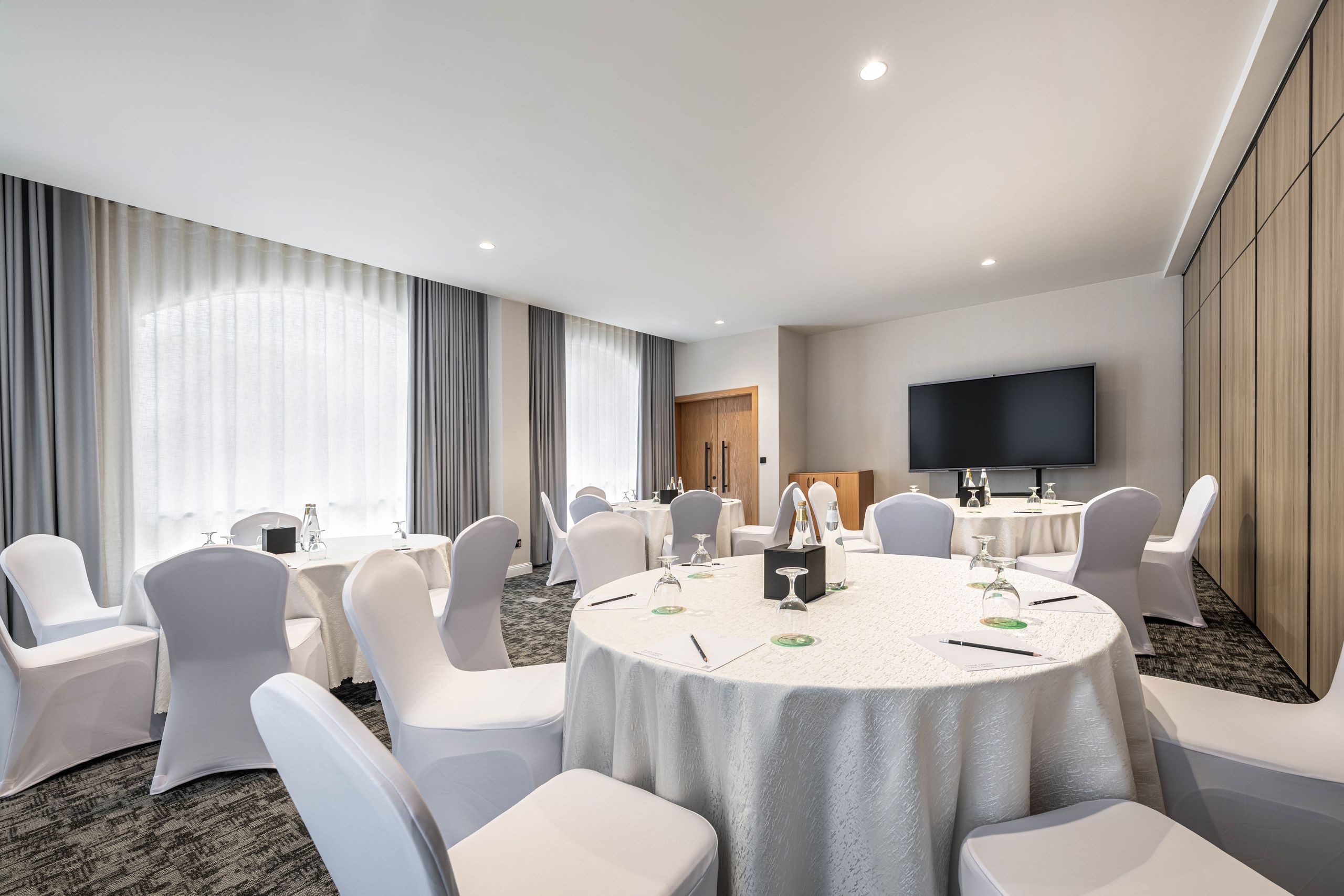 The photo shows a conference or meeting room with round tables and chairs arranged for an event.…