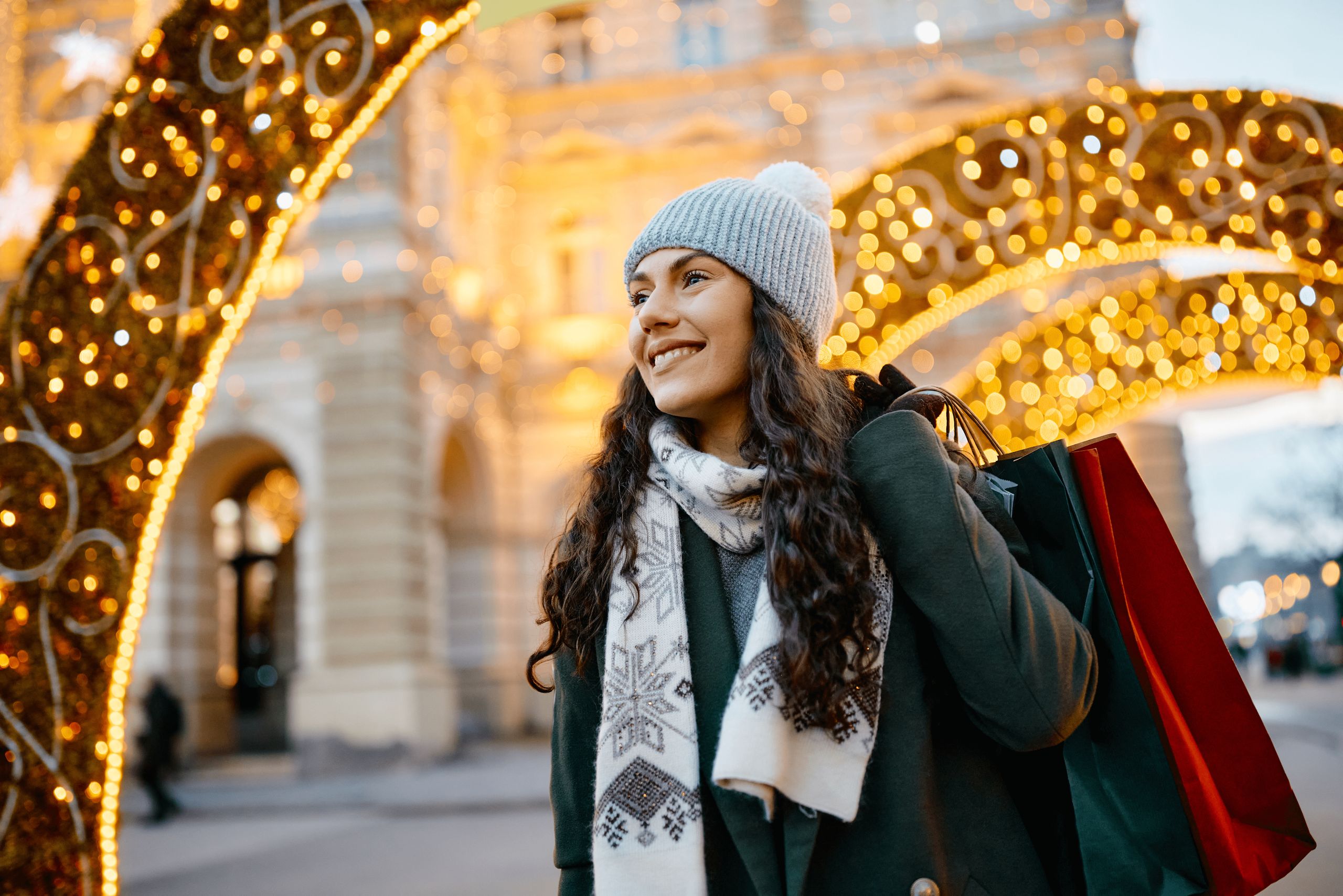 Een vrouw in winterkleding draagt boodschappentassen en staat buiten bij feestelijke verlichting.