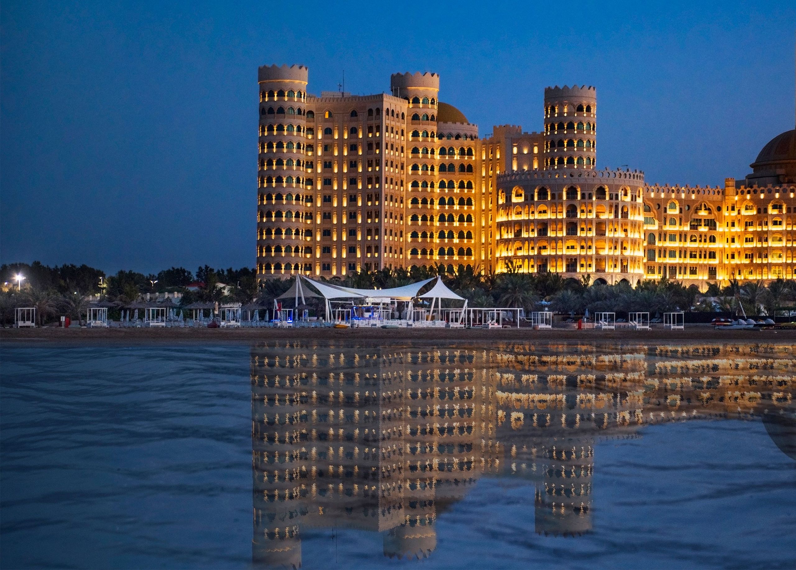 A large illuminated building by the water at dusk, with its reflection visible in the water.
