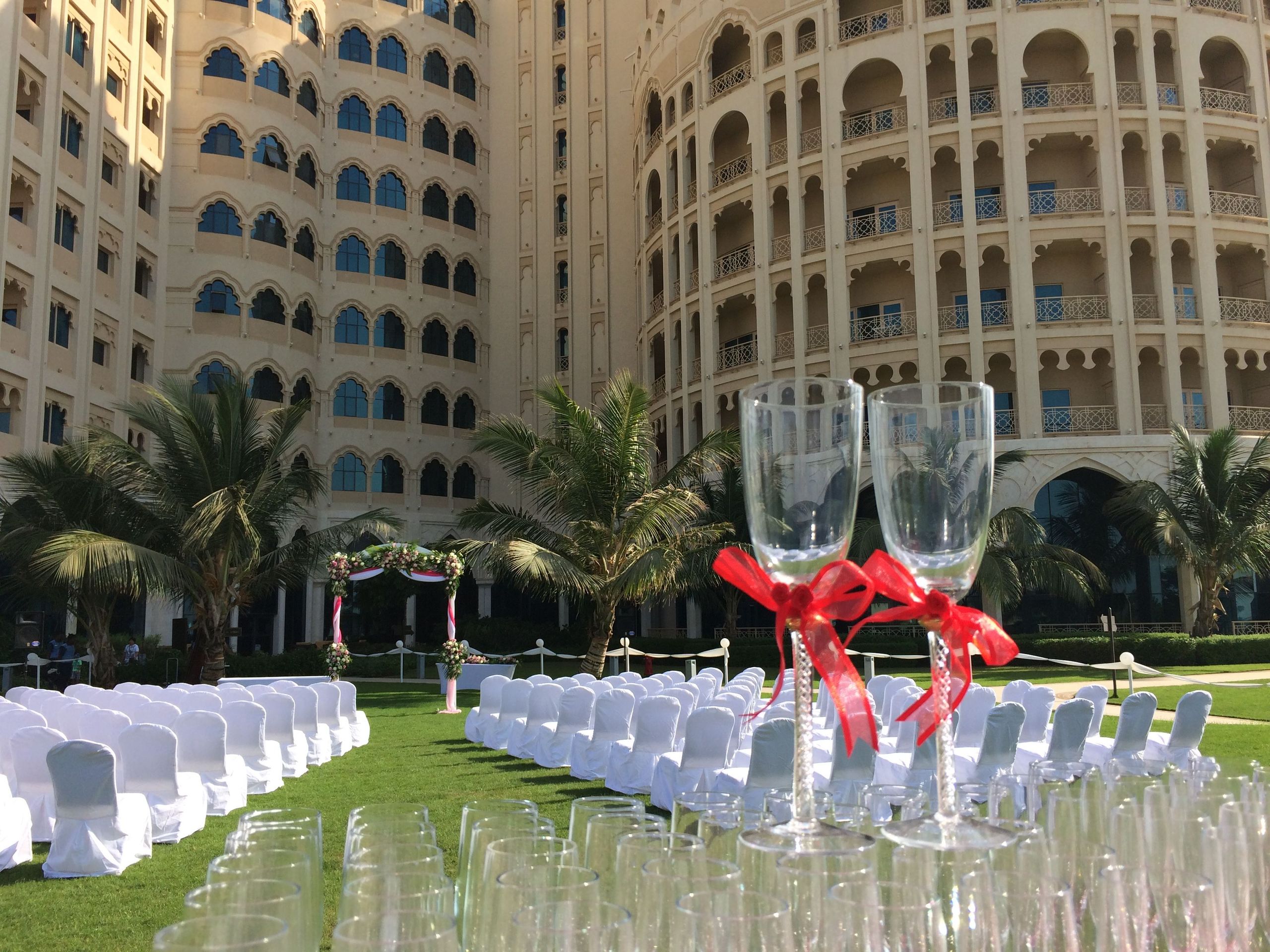 The photo shows an outdoor wedding setup with chairs arranged in rows on grass, a decorated arch,…