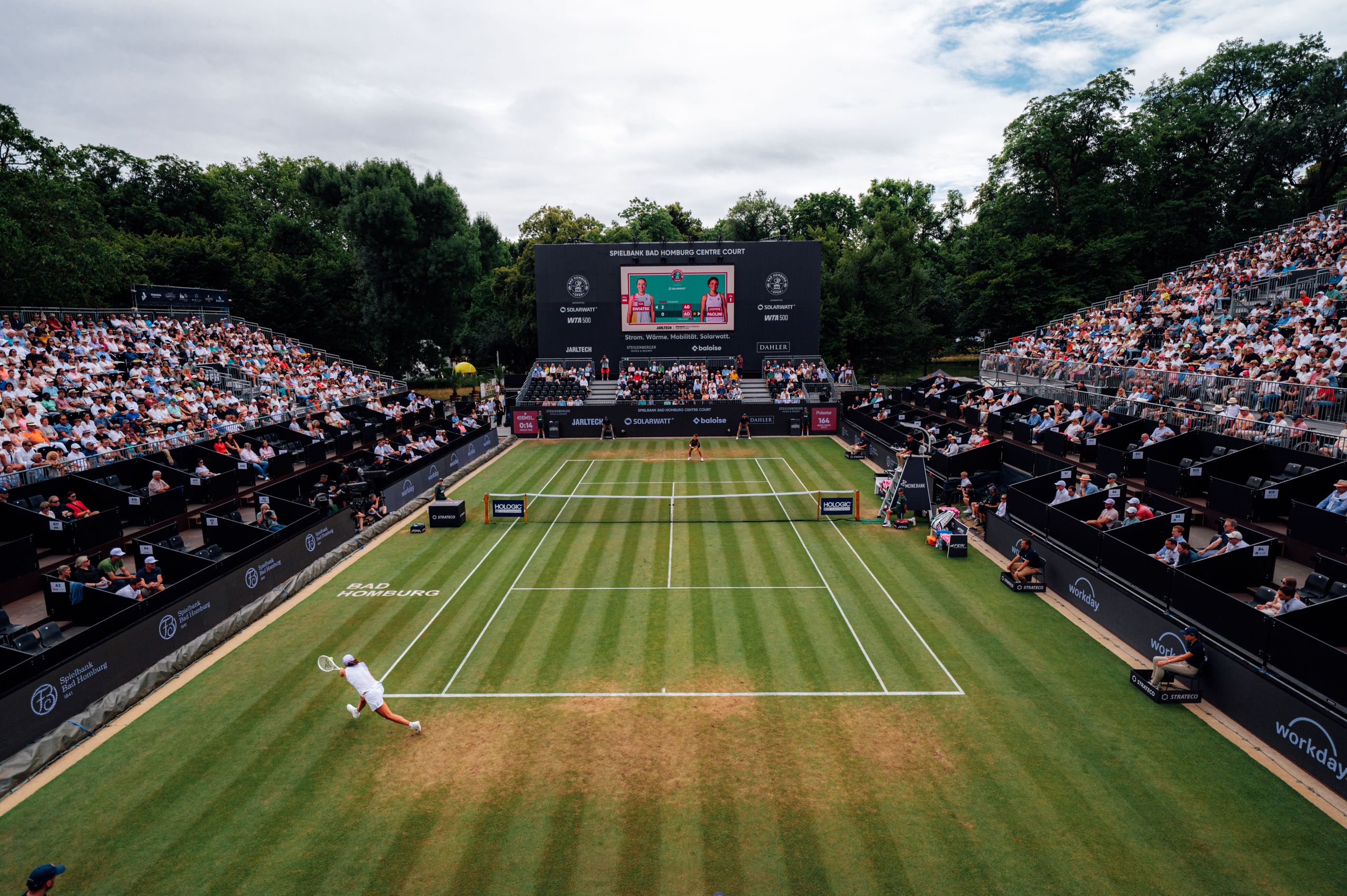A tennis match is taking place on a grass court with a large crowd of spectators watching from the…