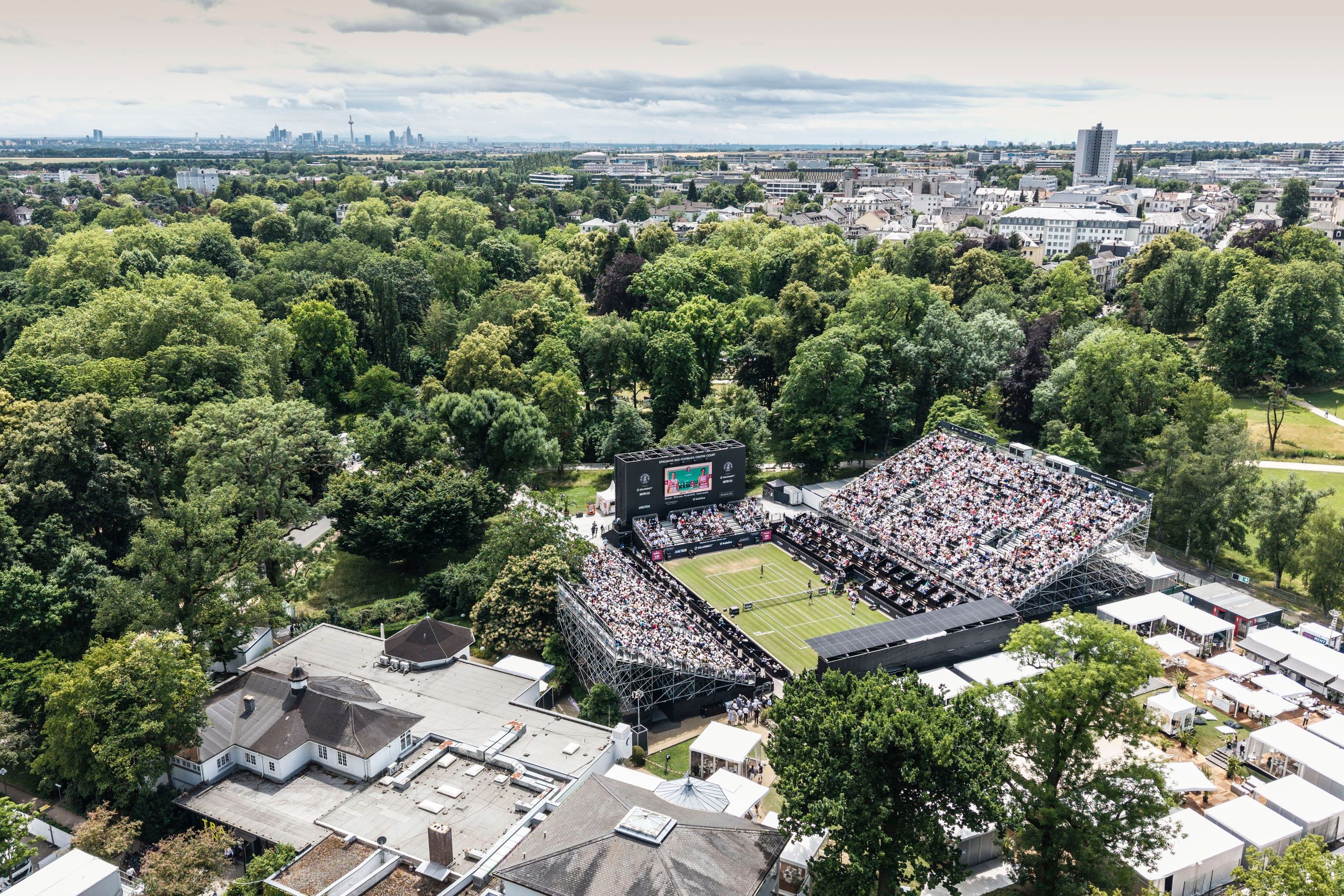 An aerial view of an outdoor tennis stadium surrounded by trees and buildings, with a tennis match…