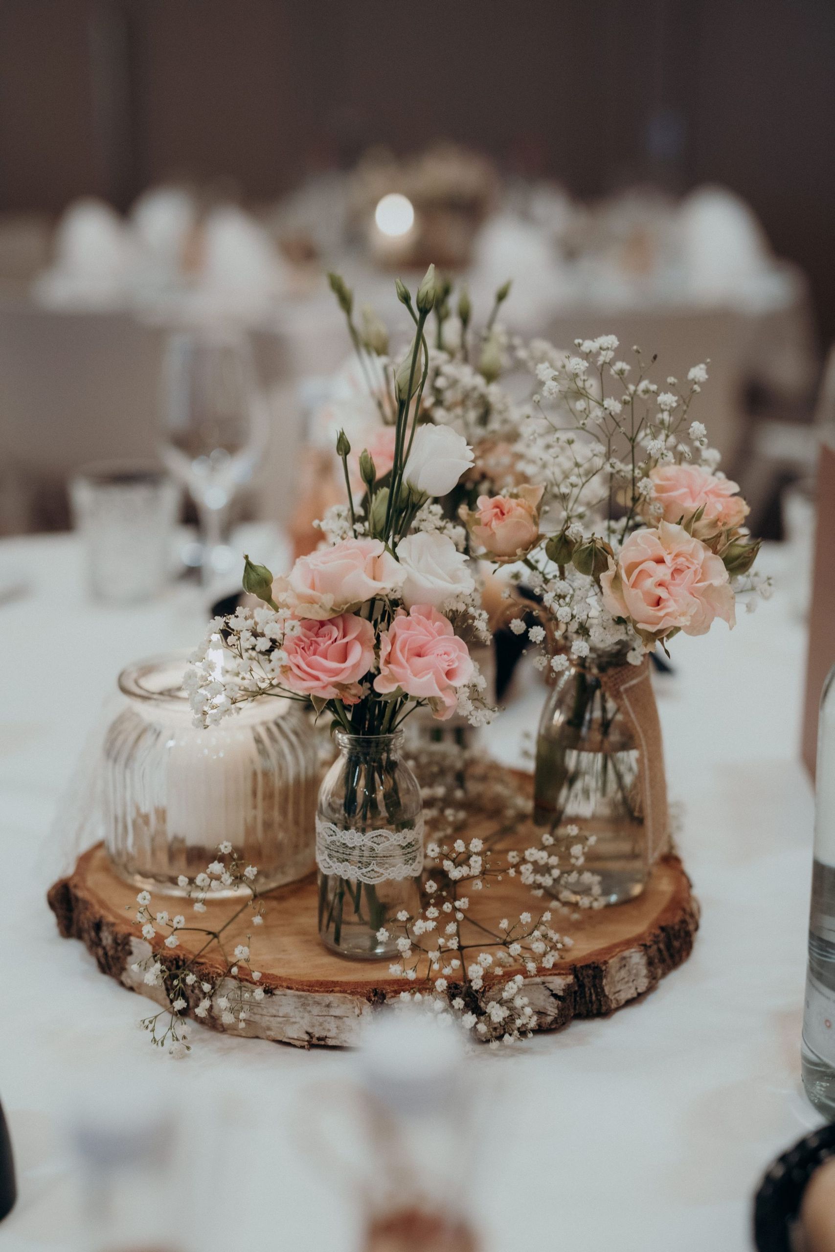 Une composition florale sur une table, comprenant de petits bocaux en verre avec des fleurs roses…