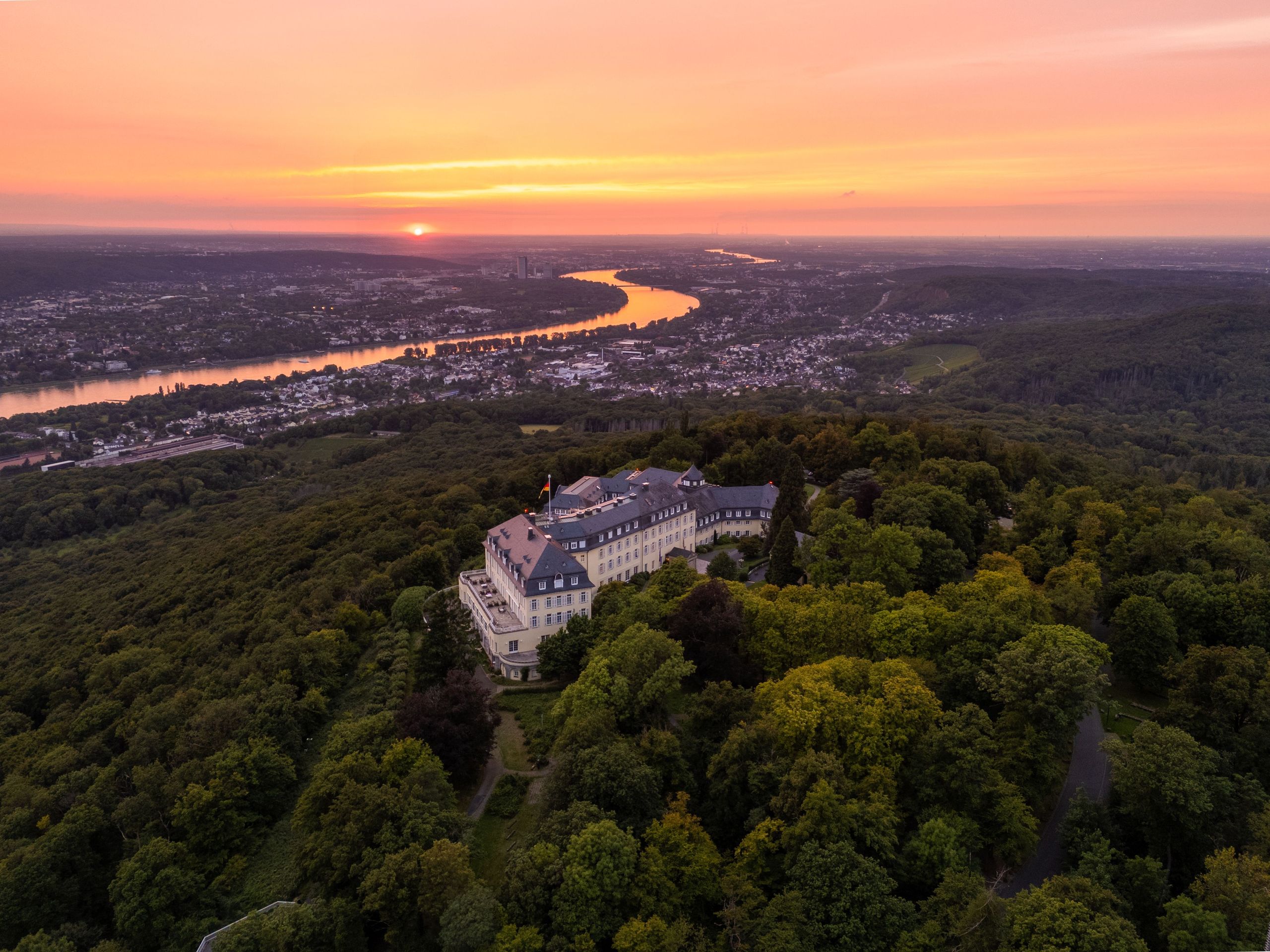 Ein großes Gebäude, umgeben von dichtem Wald, mit einem Fluss und einer Stadtlandschaft im…