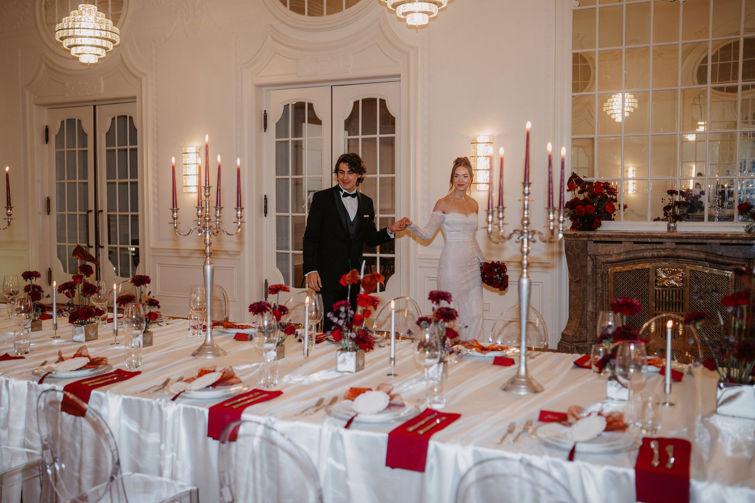 Na fotografii je formálně oblečený pár stojící v elegantně zařízené jídelně s dlouhým stolem…