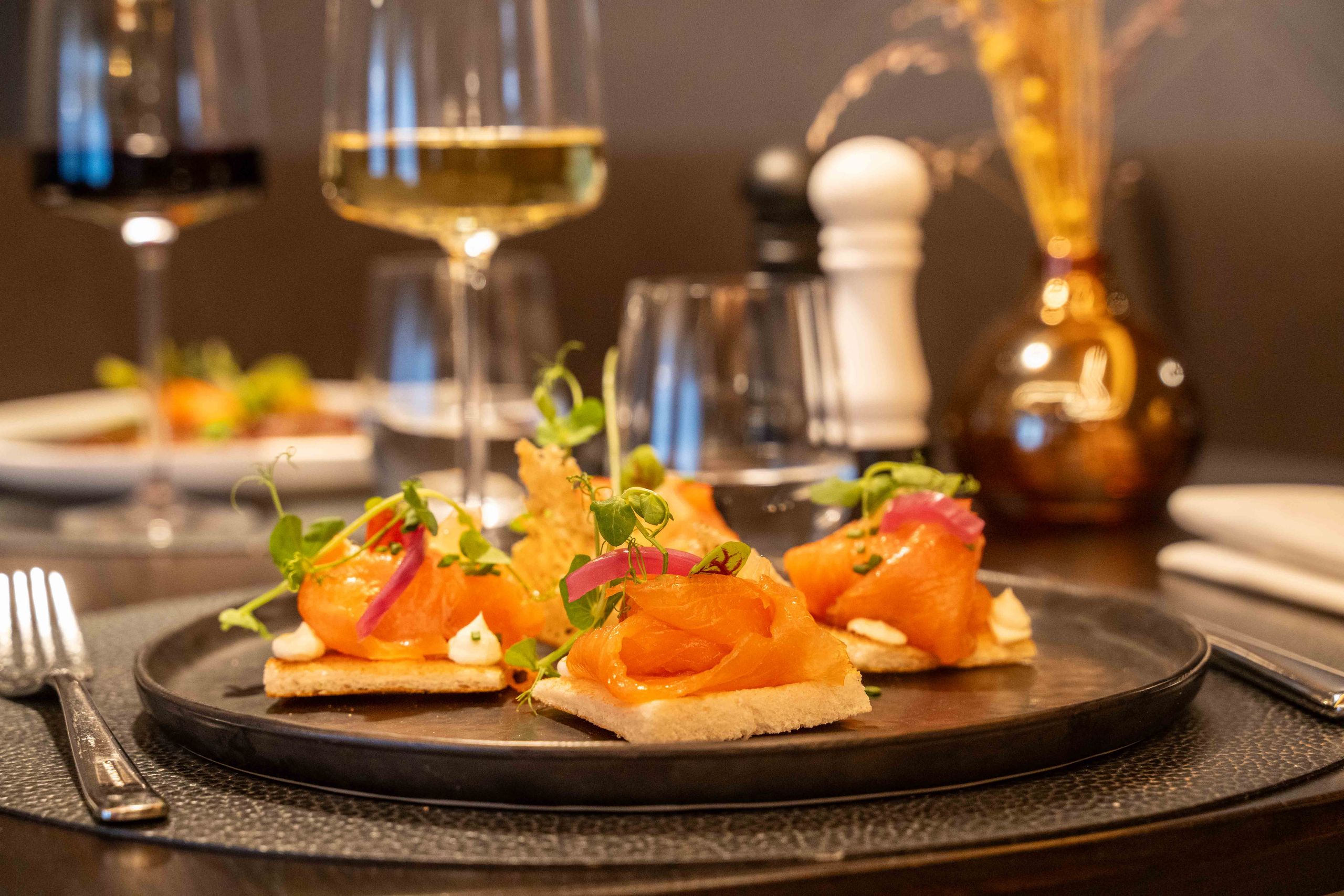 Une assiette avec des amuse-bouches au saumon fumé et des verres de vin en arrière-plan.