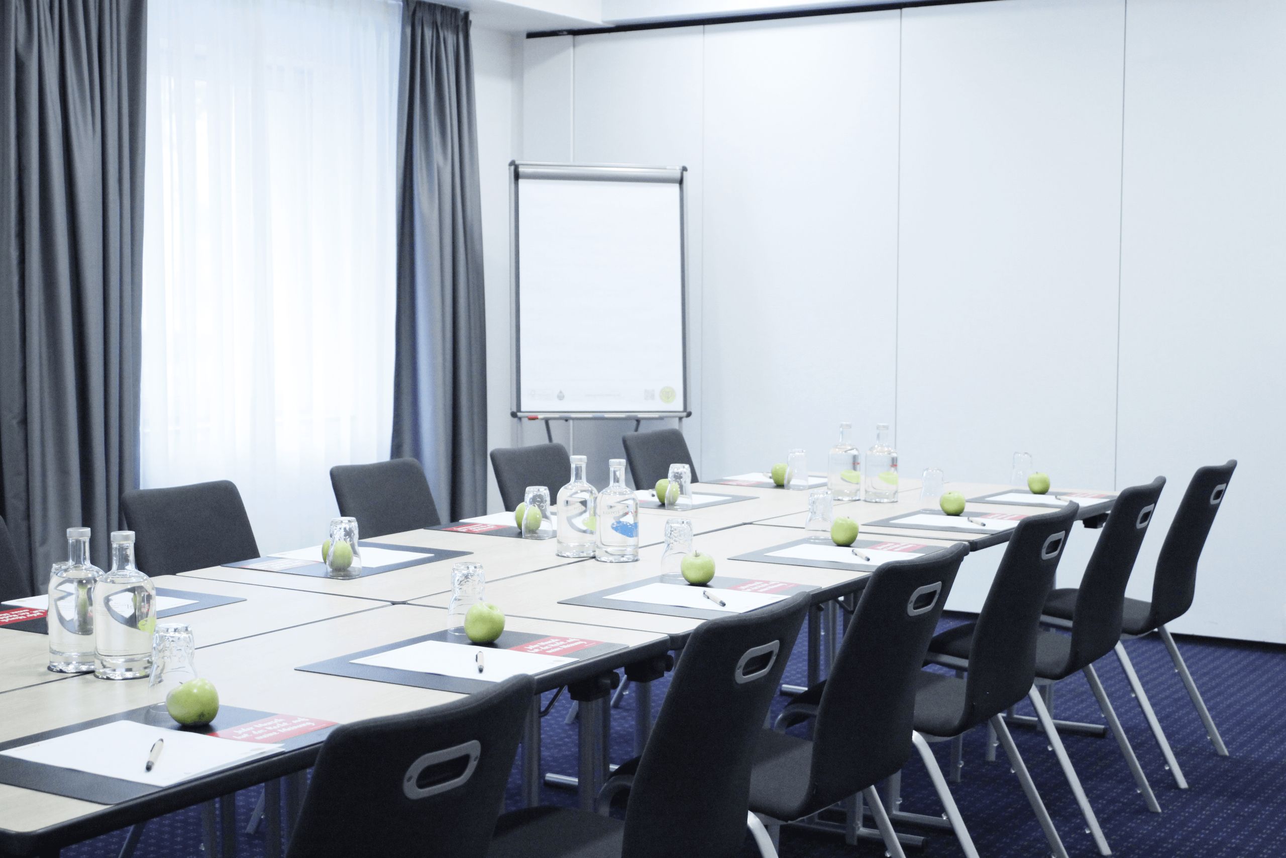 Ein Konferenzraum mit einem Tisch, Stühlen, Notizblöcken, grünen Äpfeln, Wasserflaschen und einem Flipchart.